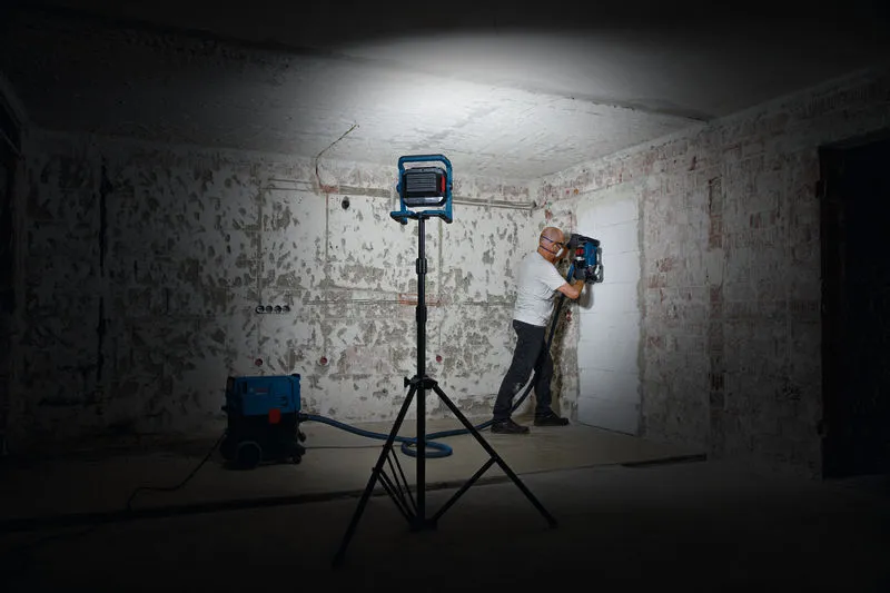 Une personne portant un équipement de sécurité utilise un outil de perçage mural sous une lampe de travail.