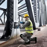 Une personne portant un équipement de sécurité utilise une perceuse à noyau magnétique sur une poutre de pont en acier.