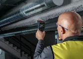 Une personne portant un équipement de sécurité utilise un détecteur thermique pour vérifier une gaine de ventilation.