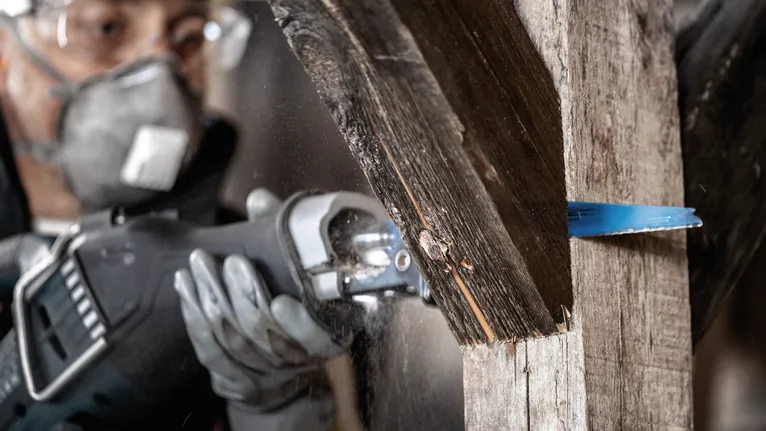 Une personne portant un équipement de sécurité utilise une scie alternative pour couper une poutre en bois.