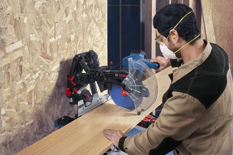 Une personne portant un équipement de sécurité utilise une scie à onglet pour couper une planche de bois.