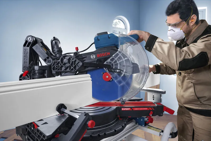 Une personne portant un équipement de sécurité utilise une scie à onglet pour couper des moulures à l'intérieur.