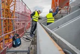 Deux personnes portant des équipements de sécurité travaillent sur un échafaudage avec des outils de construction et des coffrages en béton.