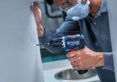 Une personne portant un équipement de sécurité perce une armoire avec une visseuse à percussion sans fil.