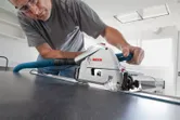 Une personne portant un équipement de sécurité coupe un panneau avec une scie plongeante fixée à un rail de guidage.