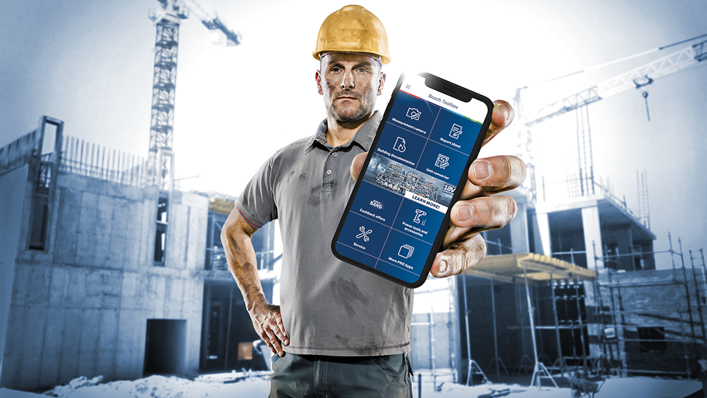 Un ouvrier en casque montre une application sur un téléphone, chantier en arrière-plan.