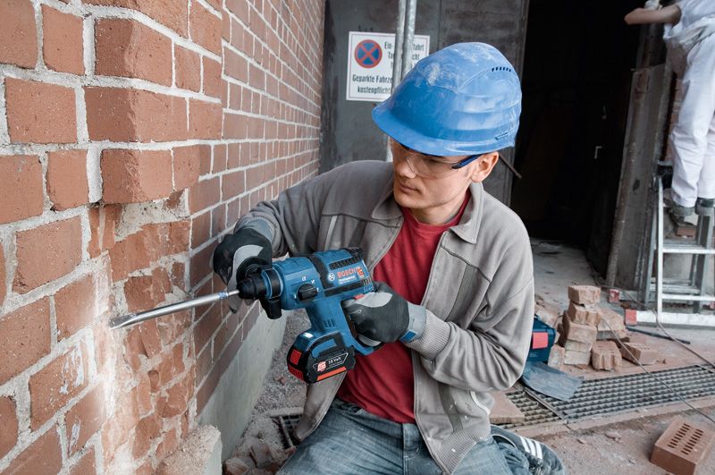 A person wearing safety equipment drills into a brick wall using a cordless rotary hammer.