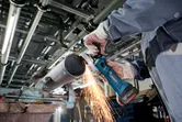 A person wearing safety equipment grinds a metal pipe with a cordless angle grinder.