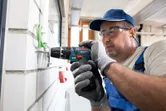 A person wearing safety equipment uses a cordless combi drill on a wall.
