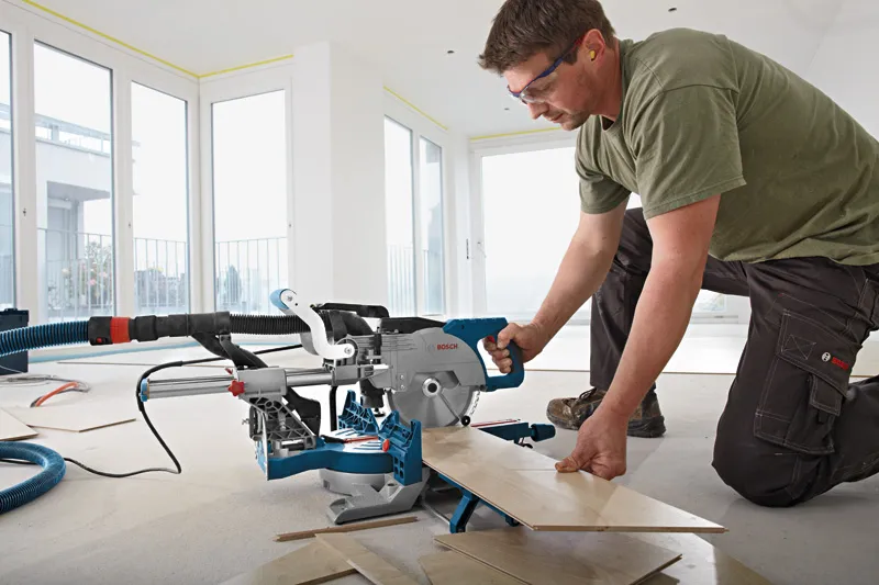 A person wearing safety equipment cuts floorboards with a sliding miter saw indoors.