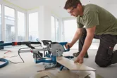 A person wearing safety equipment cuts floorboards with a sliding miter saw indoors.