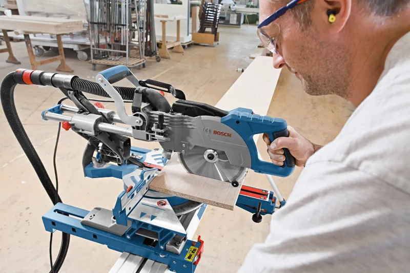 A person wearing safety equipment cuts wood with a sliding mitre saw in a workshop.