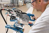 A person wearing safety equipment cuts wood with a sliding mitre saw in a workshop.