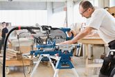 A person wearing safety equipment operates a sliding mitre saw on a workbench.