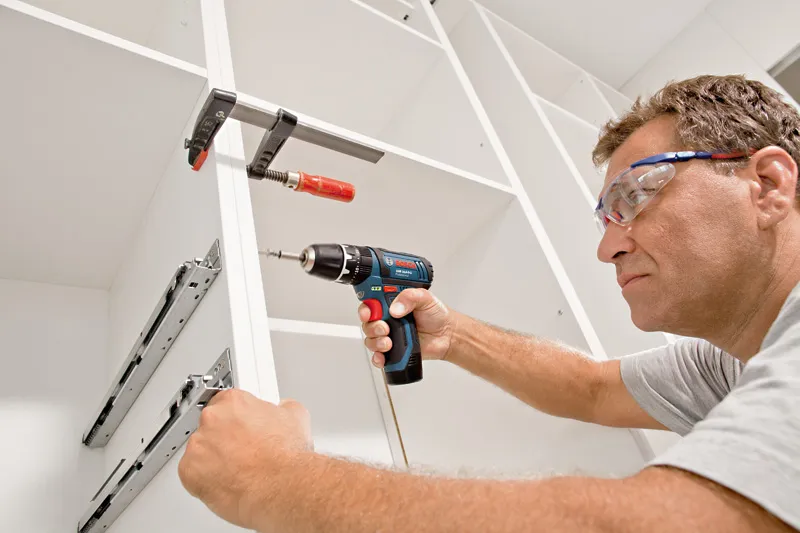 A person wearing safety equipment uses a cordless drill to assemble a cabinet.