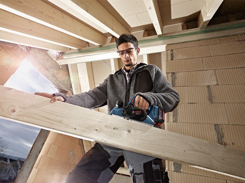 A person wearing safety equipment uses a cordless planer on a wooden beam.
