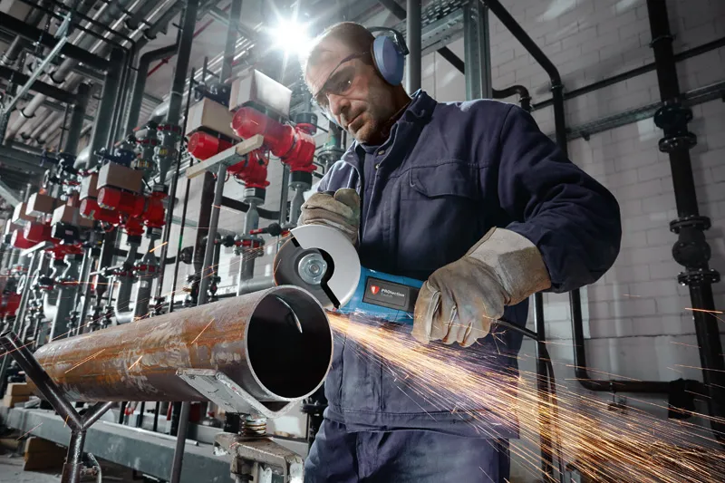 A person wearing safety equipment uses an angle grinder to cut a large metal pipe.