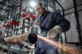 A person wearing safety equipment uses an angle grinder to cut a large metal pipe.
