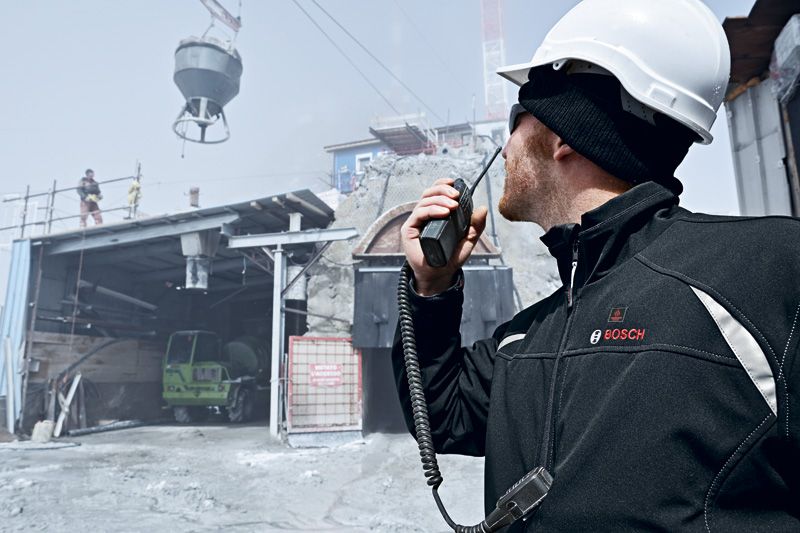 A person wearing safety equipment communicates with a radio at a busy construction site.
