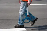 A person wearing safety equipment measures distance on asphalt with a measuring wheel.