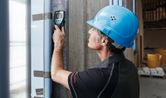 A person wearing safety equipment scans a concrete wall with a wallscanner detector.