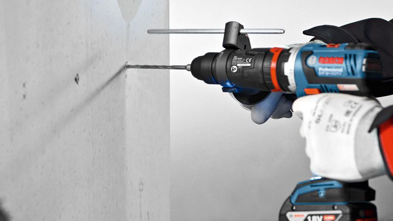 A person wearing safety equipment drills into a concrete wall with a power tool adapter.