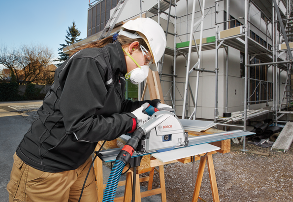 Person wearing safety equipment operates a track saw to cut sheet material at a construction site.