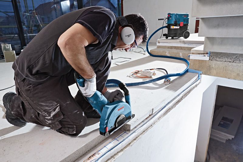 A person wearing safety equipment uses a wall chaser on concrete near a vacuum system.