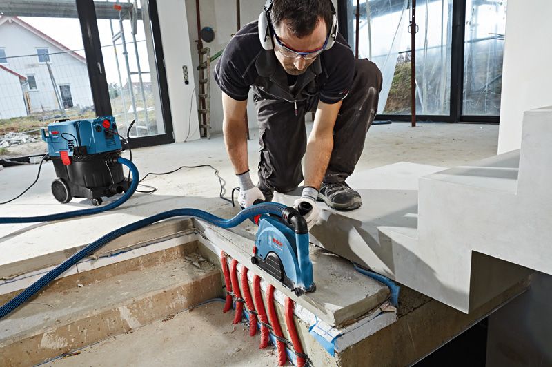 A person wearing safety equipment uses a wall chaser on concrete above exposed pipes.