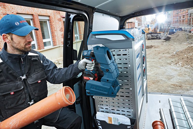 A person wearing safety equipment holds a cordless tool inside a work van at a construction site.