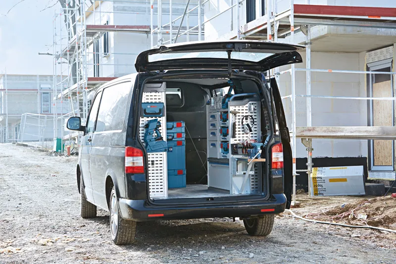 Open van with organized storage racks and blue toolboxes parked at a construction site.