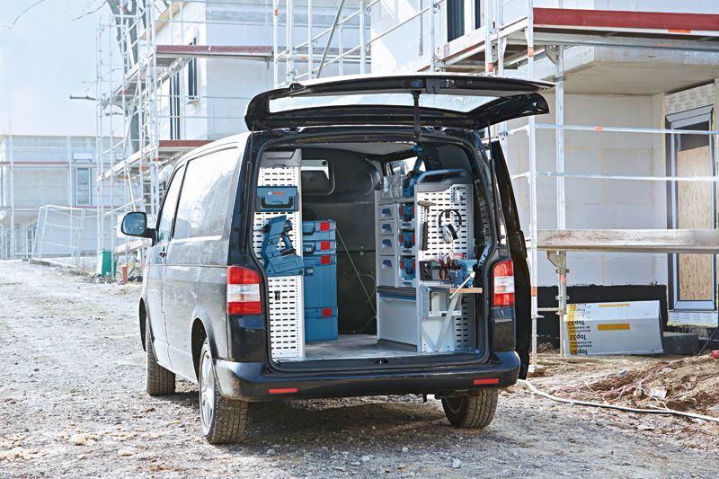 Open van with organized storage racks and blue toolboxes parked at a construction site.