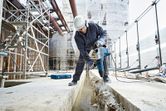 A person wearing safety equipment uses a rotary hammer to break concrete at a construction site.