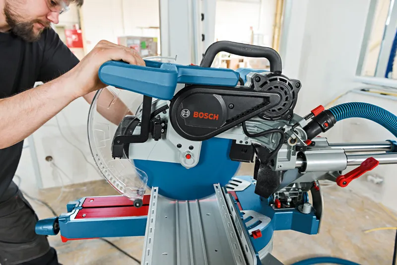 A person wearing safety equipment operates a sliding mitre saw for precise wood cutting.