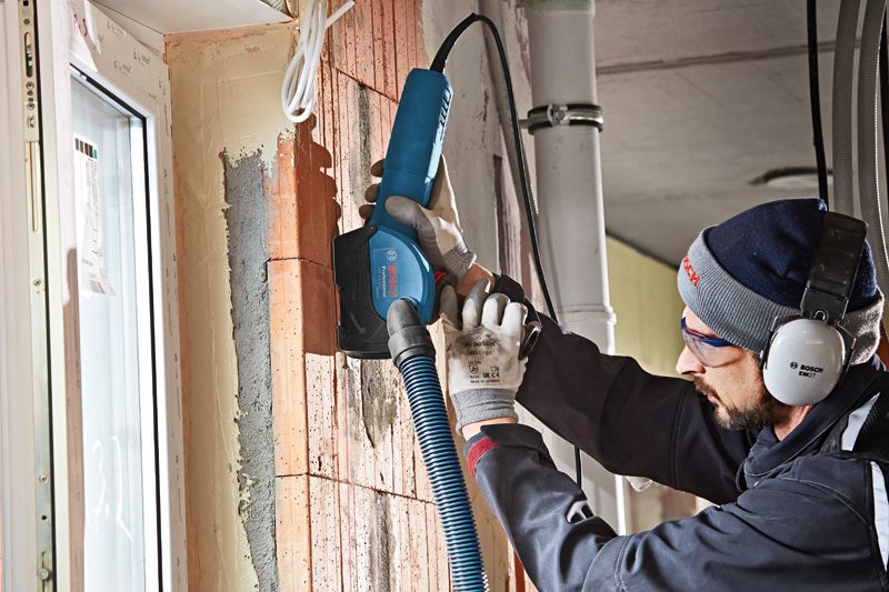 A person wearing safety equipment uses a wall chaser with a dust extraction hose on brick.