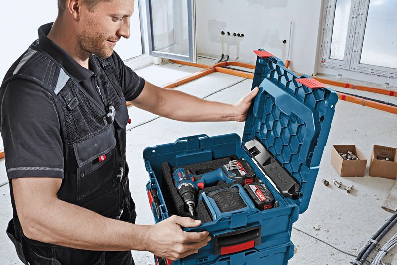 A person wearing safety equipment opens a tool case containing a cordless drill and accessories.