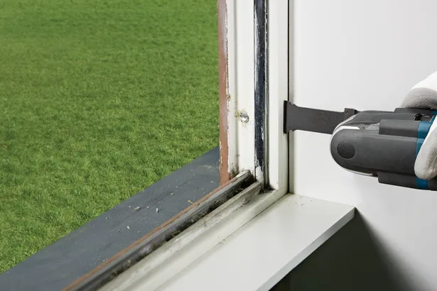Gloved hand uses a multitool to cut sealant near a window frame.