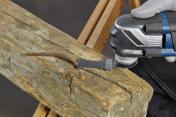 Worker wearing safety equipment cuts through a rusty nail in a wooden beam with a multi-tool.