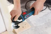 A person cuts wood at the floor with a multi-cutter power tool.