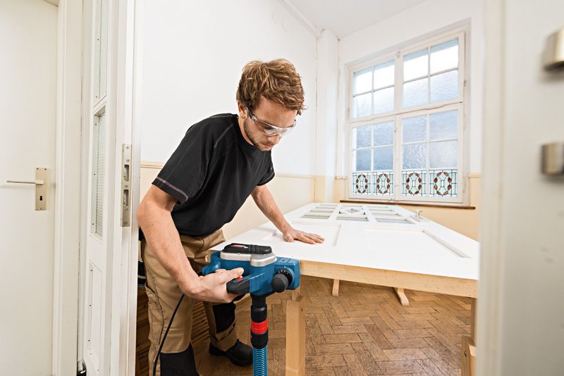 A person wearing safety equipment uses a planer on a door in a bright room.