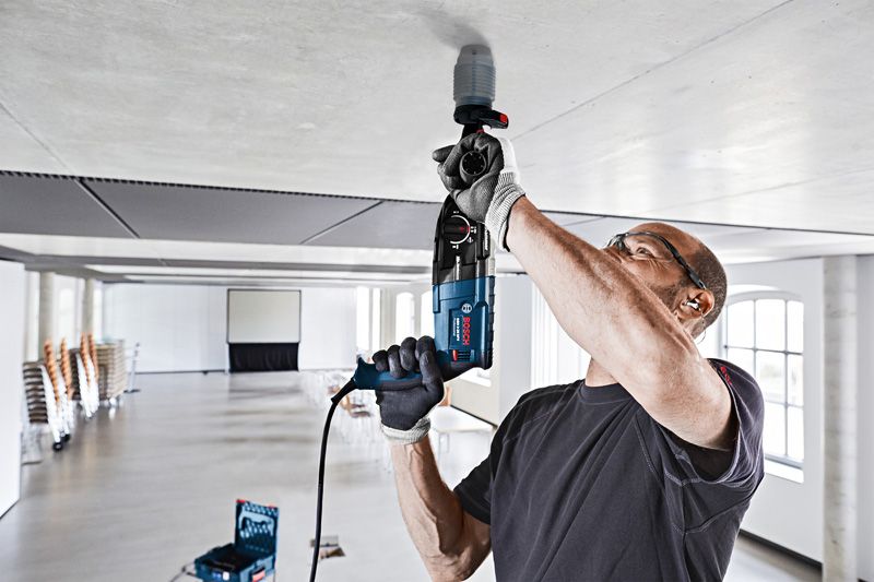 A person wearing safety equipment drills into a ceiling with a dust collection cover attached.