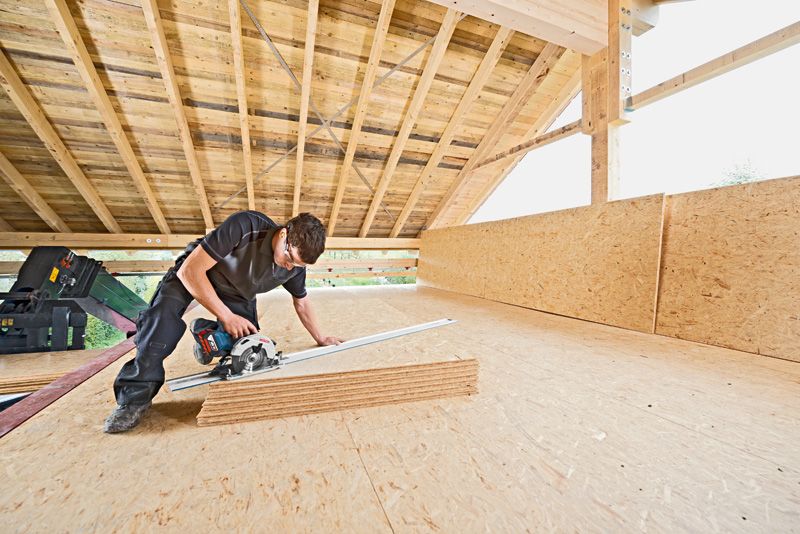 A person wearing safety equipment cuts plywood sheets with a cordless circular saw.