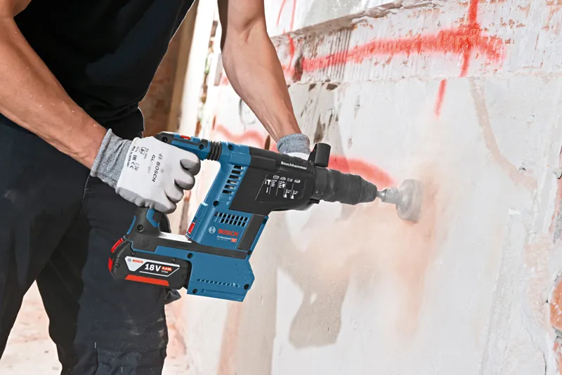 A person wearing safety equipment drills into a concrete wall with a cordless rotary hammer.