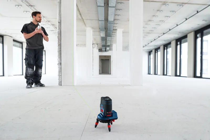 A person stands in a large empty room while a laser leveling tool accessory projects a green line.
