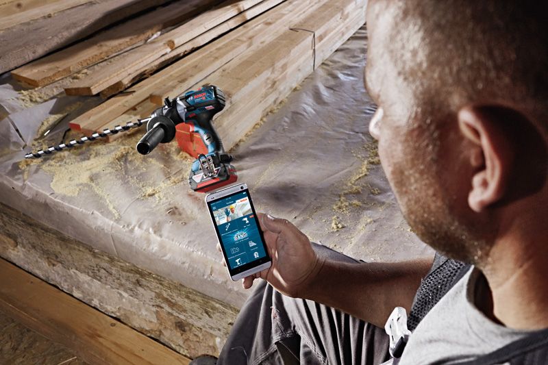 A person uses a smartphone to control a cordless drill driver on a construction site.