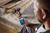 A person uses a smartphone to control a cordless drill driver on a construction site.