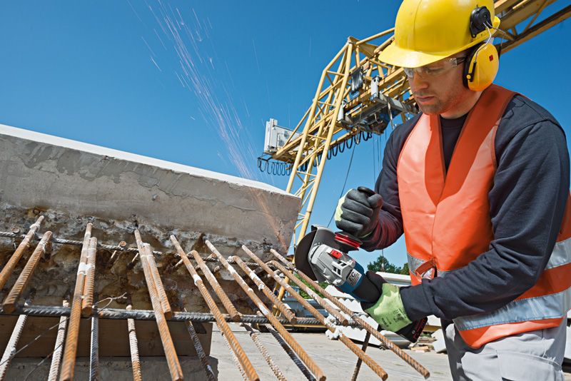 A person wearing safety equipment cuts rebar on a construction site with a cordless angle grinder.
