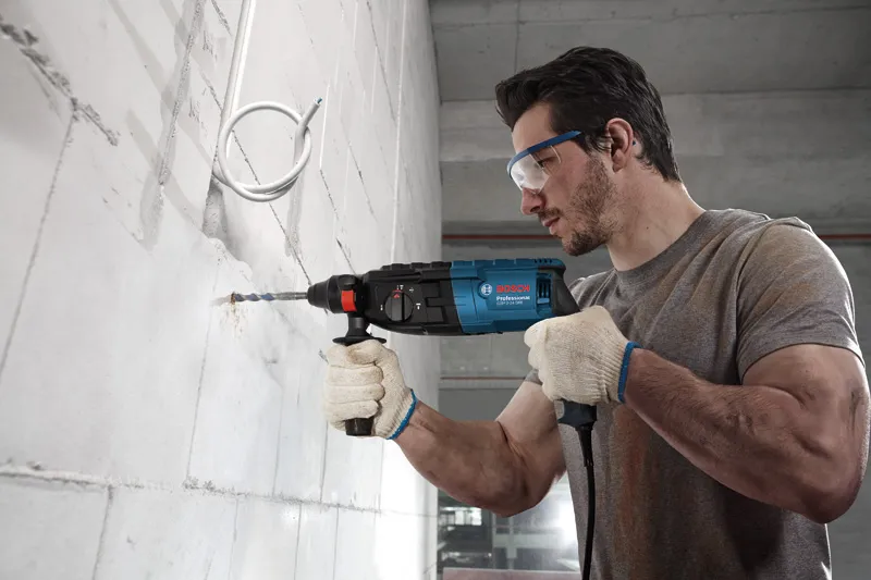 A person wearing safety equipment drills into a concrete wall with a power tool.