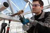 A person wearing safety equipment cuts a metal pipe with a reciprocating saw.
