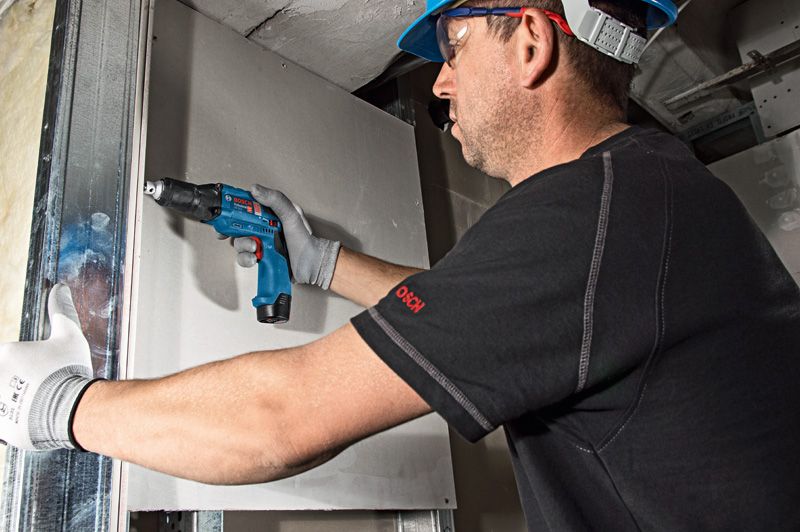 A person wearing safety equipment drives screws into drywall with a cordless screwdriver.